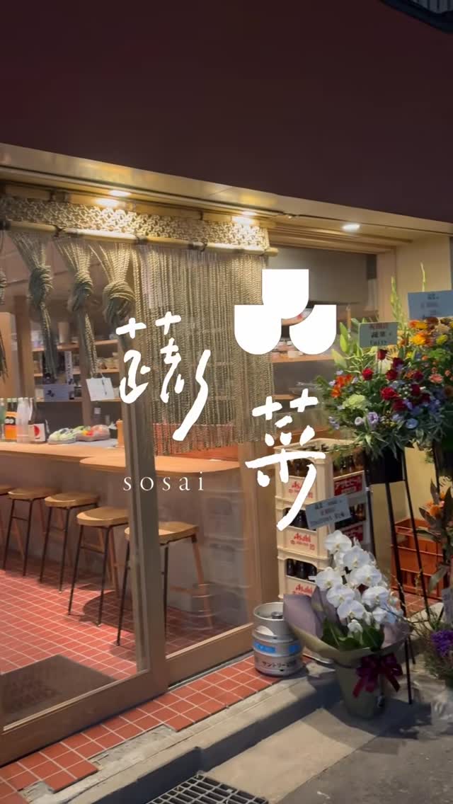 開店祝いの花が並ぶ店先。ガラス越しに木のカウンターとスツールが見える。